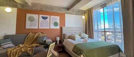 Una camera d'albergo moderna con un letto, una poltrona, un tavolino e un'ampia finestra che si affaccia sulla città.