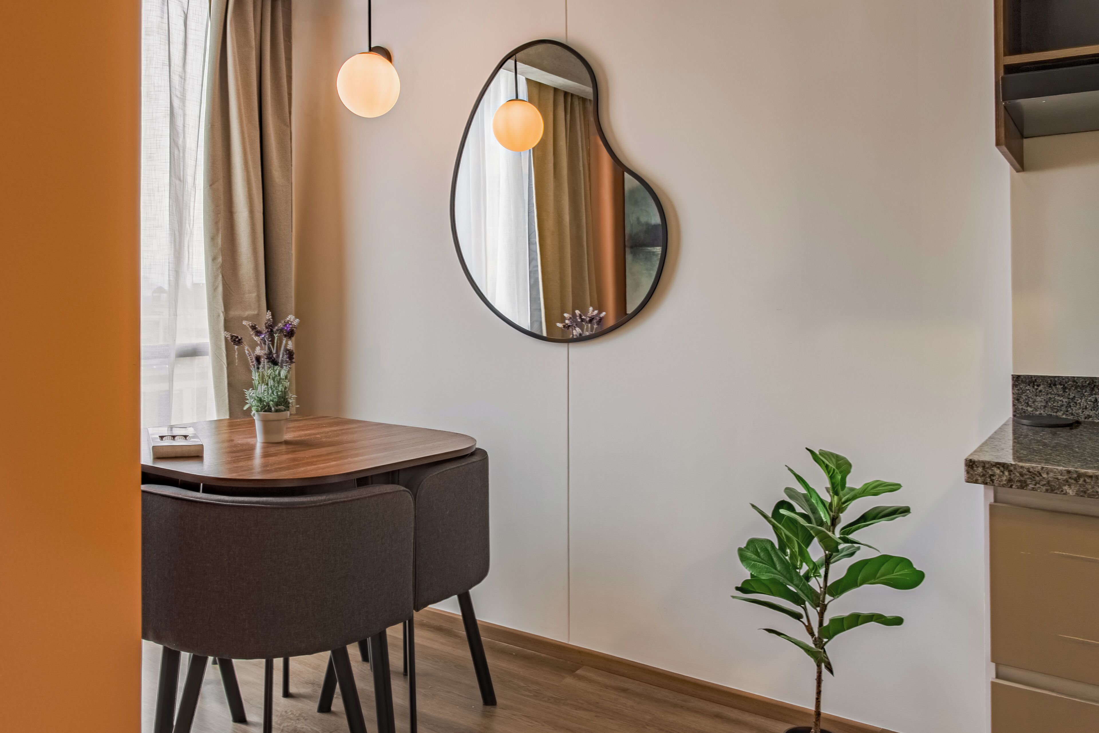 A modern dining area with a round table, two chairs, a potted plant, a unique wall mirror, and a kitchen counter.