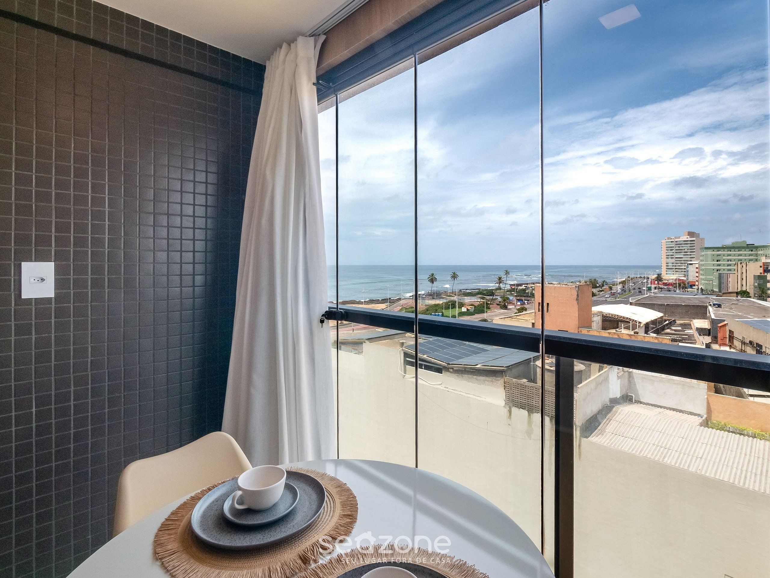 A balcony with a view of the city and ocean, a table with a cup and saucer, and a tiled wall.