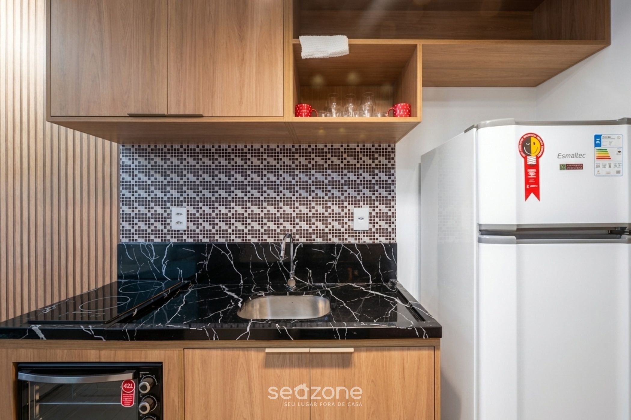 Una cucina con un piano di lavoro in marmo nero, mobili in legno, zoccolo di piastrelle, lavello in acciaio inossidabile, forno integrato e frigorifero bianco con logo Esmallec rosso.