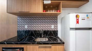 A kitchen with a black marble countertop, wooden cabinets, a tiled backsplash, a stainless steel sink, a built-in oven, and a white refrigerator with a red Esmallec logo.