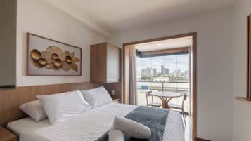 Quarto de hotel com cama, vista da cidade através de uma grande janela e uma pequena varanda com mesa e cadeiras.