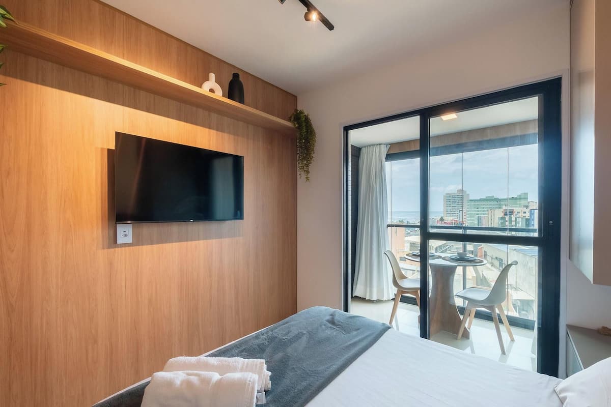 A modern hotel room with a bed, a flat-screen TV, a balcony with a table and chairs, and a city view.