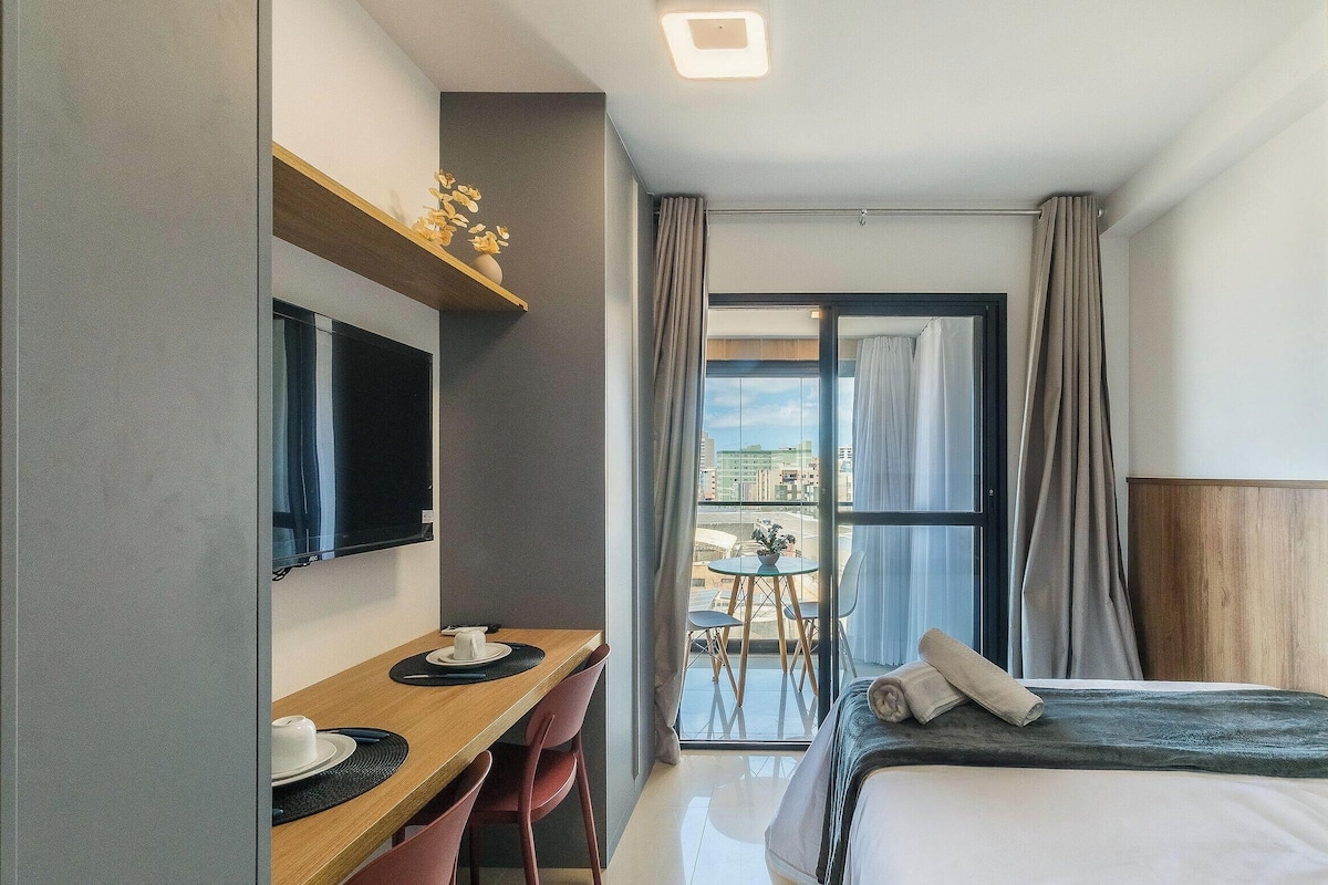 A hotel room with a bed, a desk with a chair, a TV, and a balcony with a table and chairs.