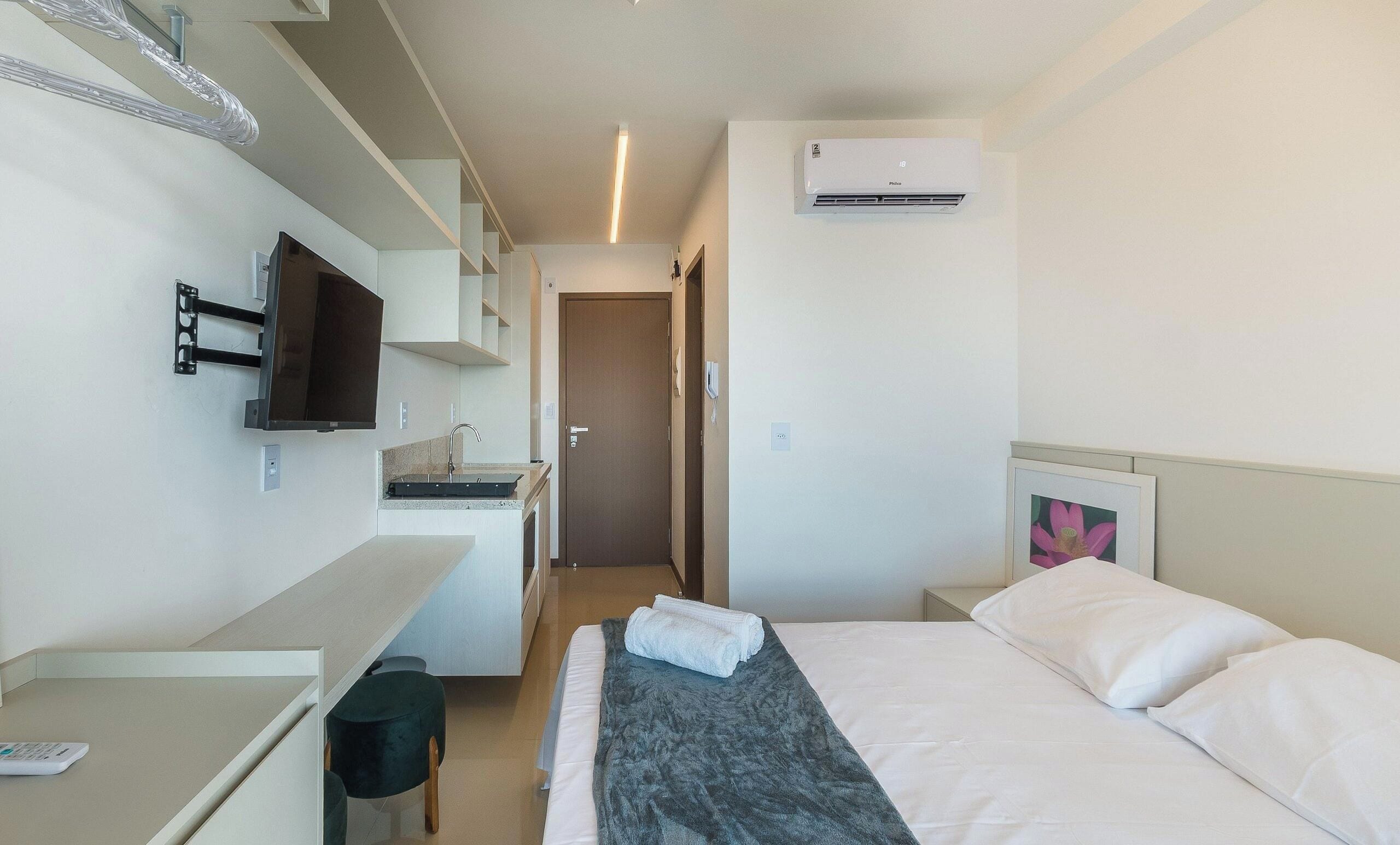 A hotel room with a bed, a desk with a flat-screen TV, a kitchenette, and an air conditioning unit.