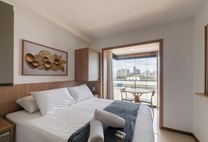 A hotel room with a bed, a view of the city, a balcony with a table and chairs, and a decorative wall piece. - Studios in Building w/ Pool and Gym - Jdns (Salvador)