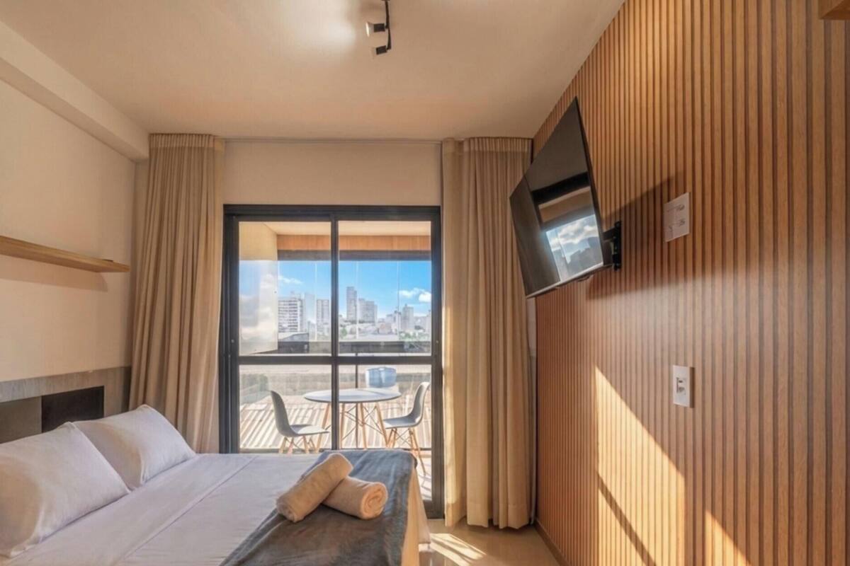 A hotel room with a bed, curtains, a TV, and a balcony with a city view.