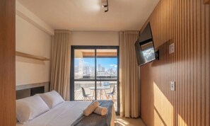 A hotel room with a bed, a TV mounted on the wall, and a balcony with a city view. - Studios in Building w/ Pool and Gym - Jdns (Salvador)