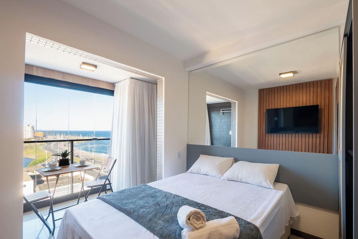 A hotel room with a bed, a TV mounted on the wall, a balcony with a table and chairs, and a view of the cityscape.
