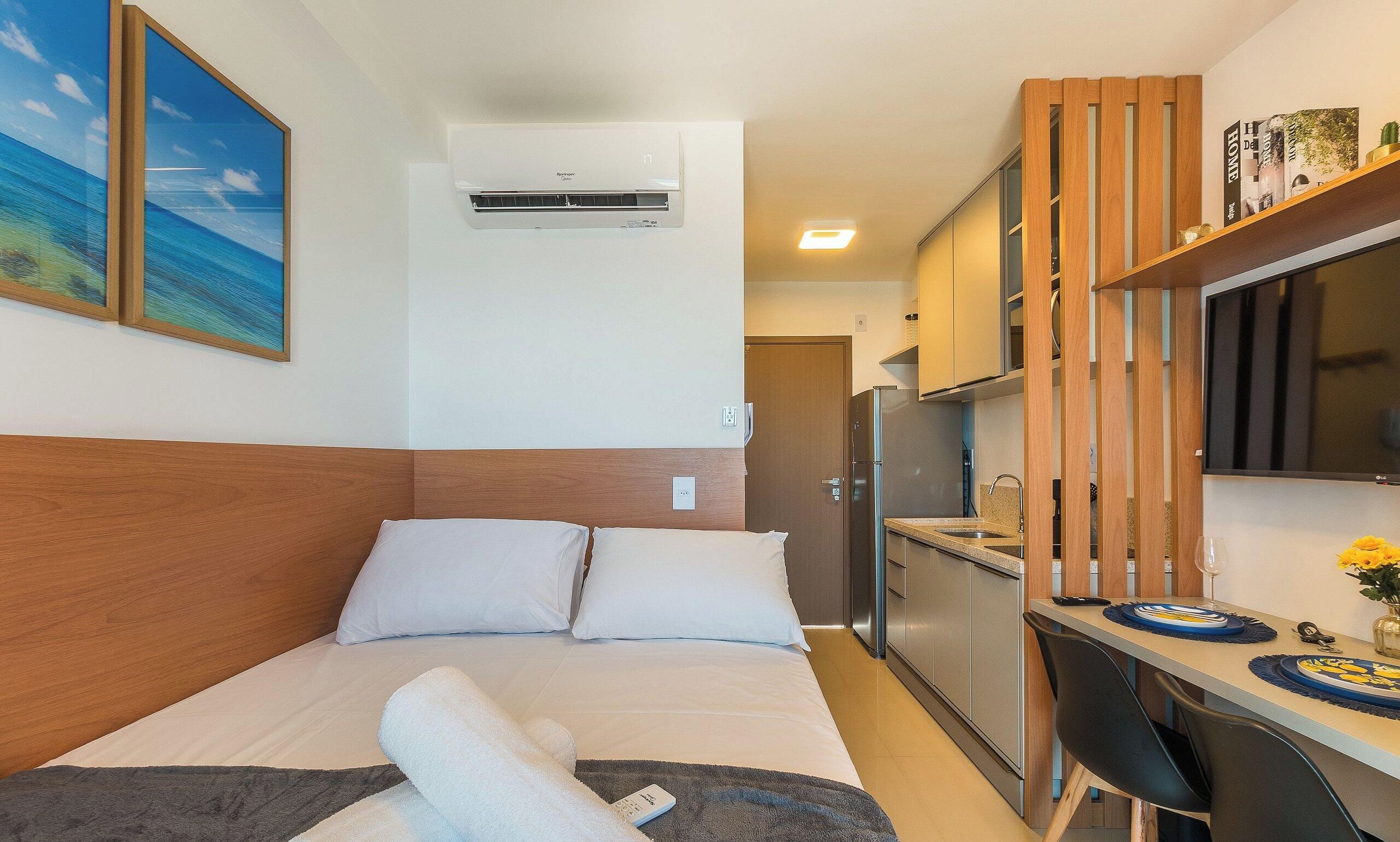 A hotel room with a bed, a desk, a refrigerator, and a wall-mounted air conditioner.
