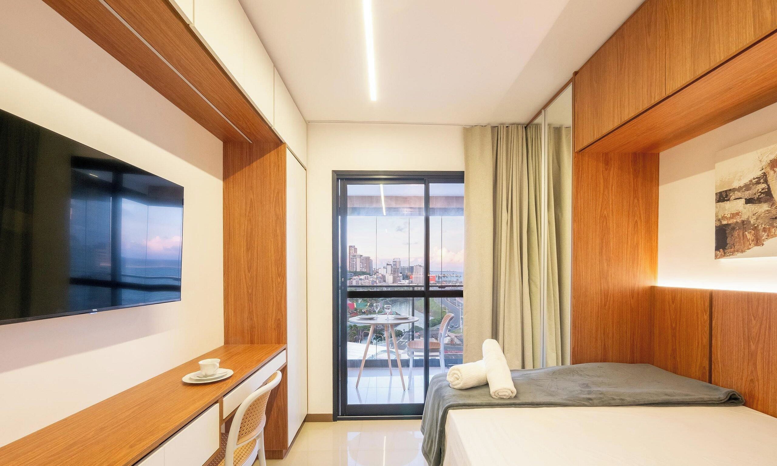 A hotel room with a bed, a desk with a chair, a television, and a window with a view of the city.
