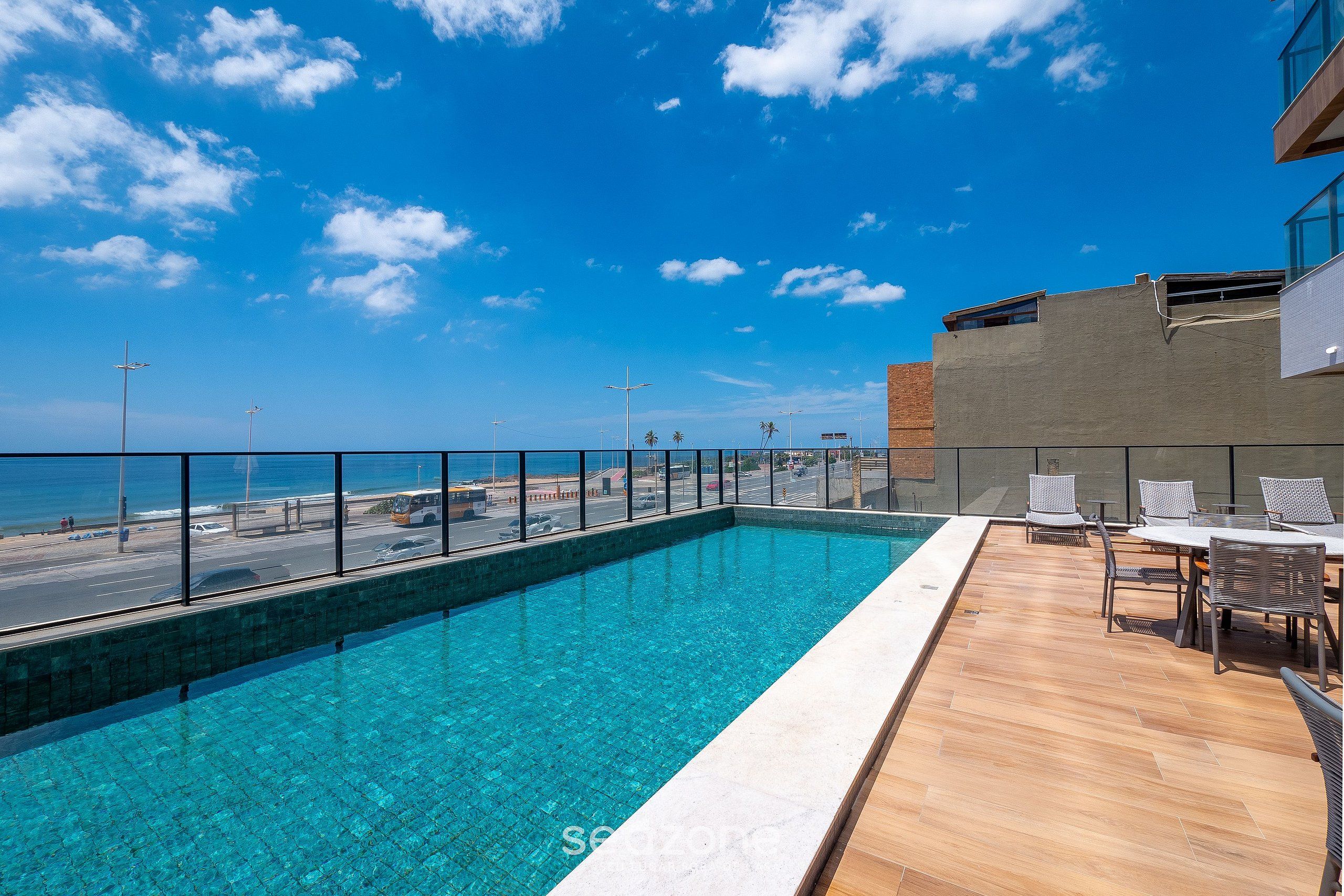 A rooftop pool with a clear view of the ocean, a wooden deck, and a seating area with chairs.