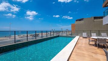 A rooftop pool with a clear view of the ocean, a wooden deck, and a seating area with chairs.