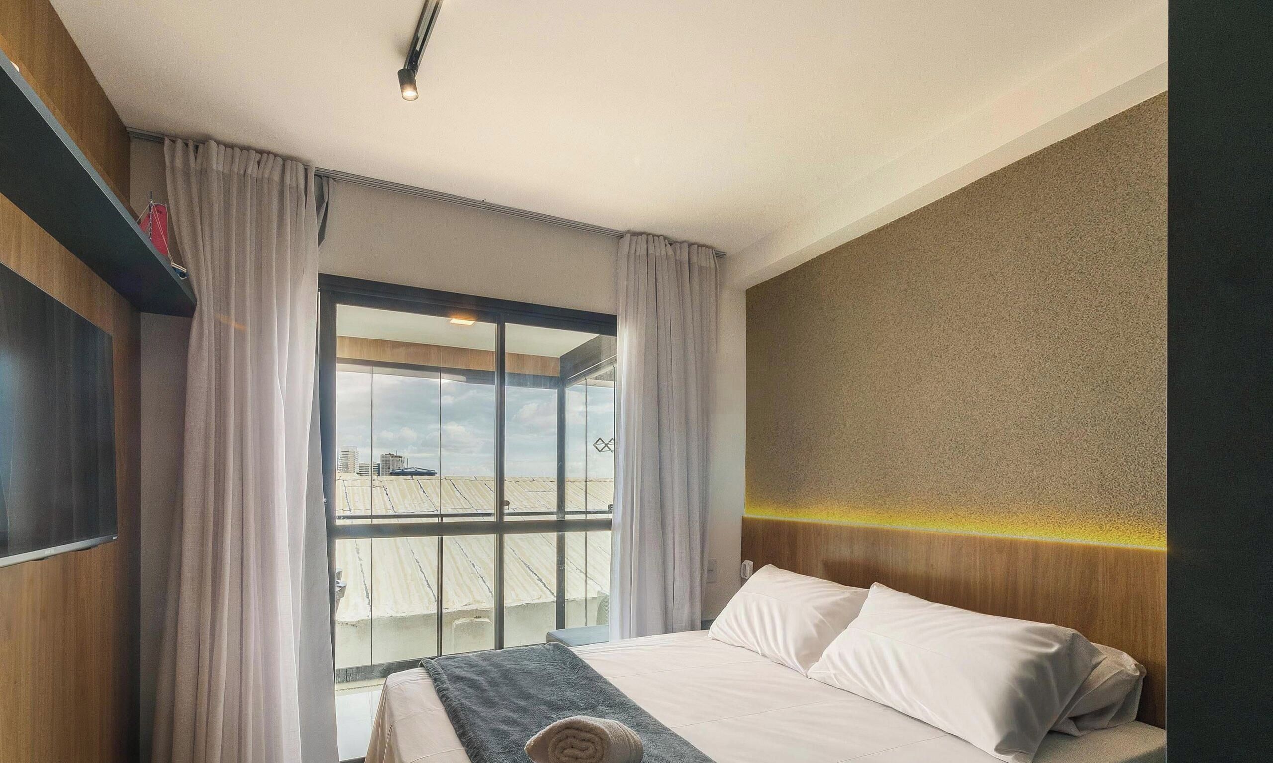 A hotel room with a bed, curtains, a TV, and a large window with a view of buildings.