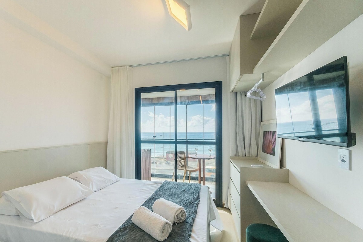A hotel room with a bed, a desk, a television, and a balcony with a view of the sea.