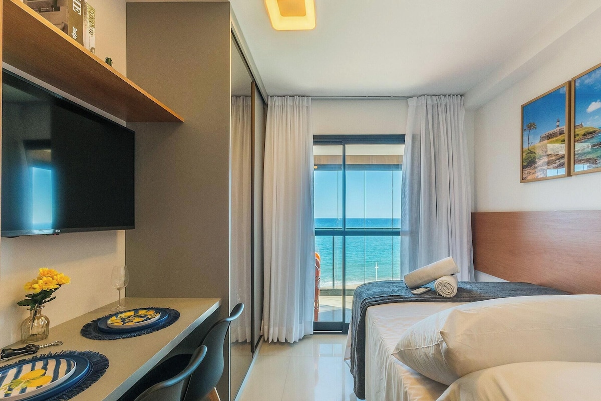 A hotel room with a bed, a desk with a flat-screen TV, a dining area, and a view of the ocean.