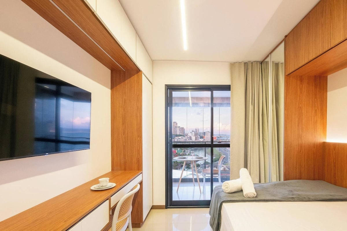 A hotel room with a bed, a desk, a TV, a balcony with a city view, and a painting on the wall.
