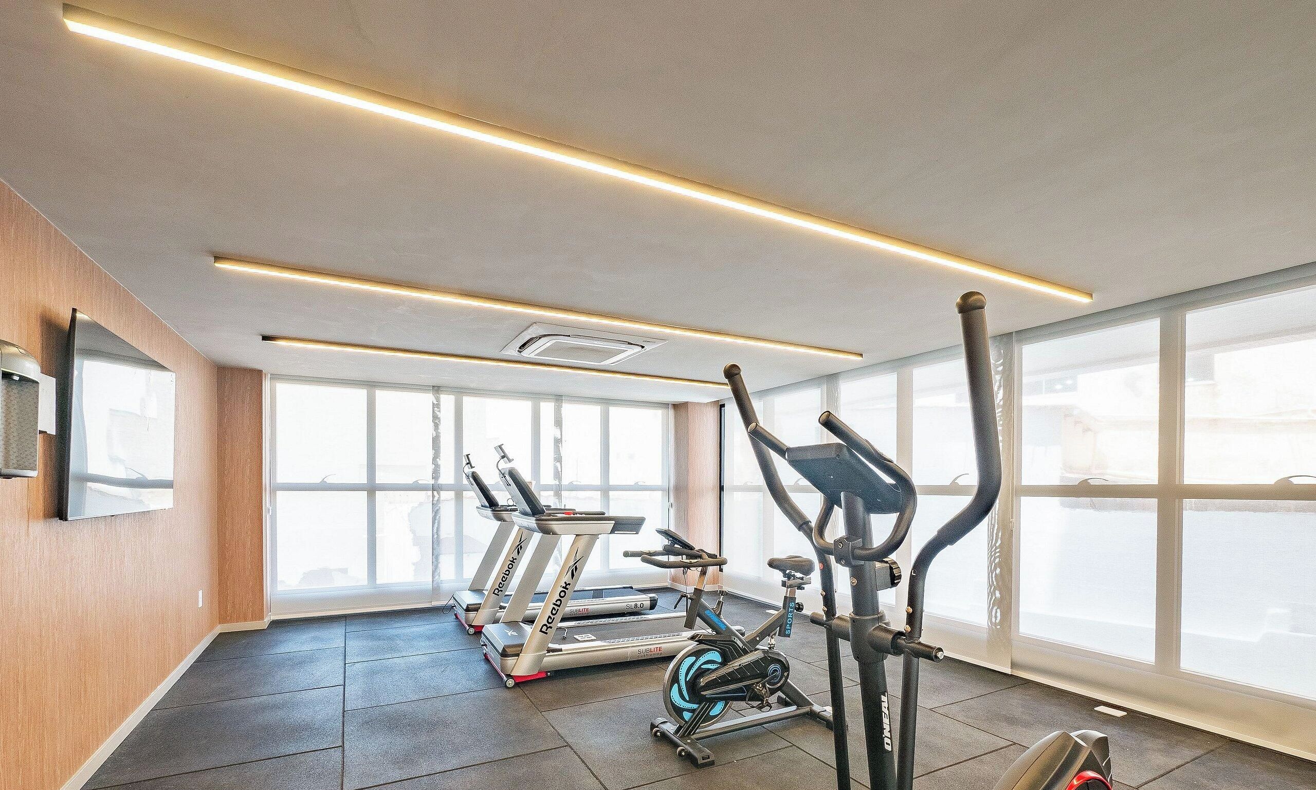 Uma academia com equipamentos de ginástica, janelas amplas e um teto com iluminação linear.