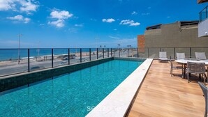 Une piscine sur le toit offrant une vue dégagée sur l’océan, une terrasse en bois et un espace détente avec des chaises.
