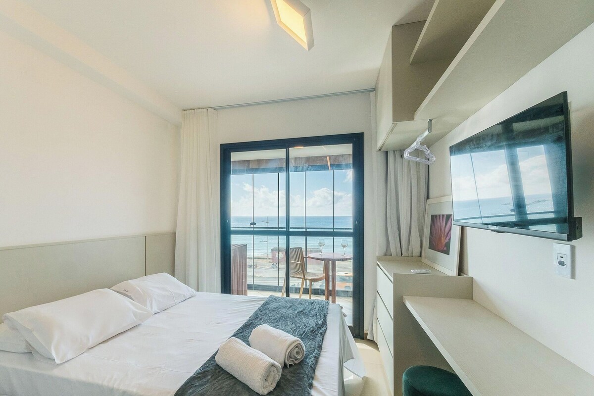 A hotel room with a bed, a desk, a chair, and a large window offering a view of the sea.