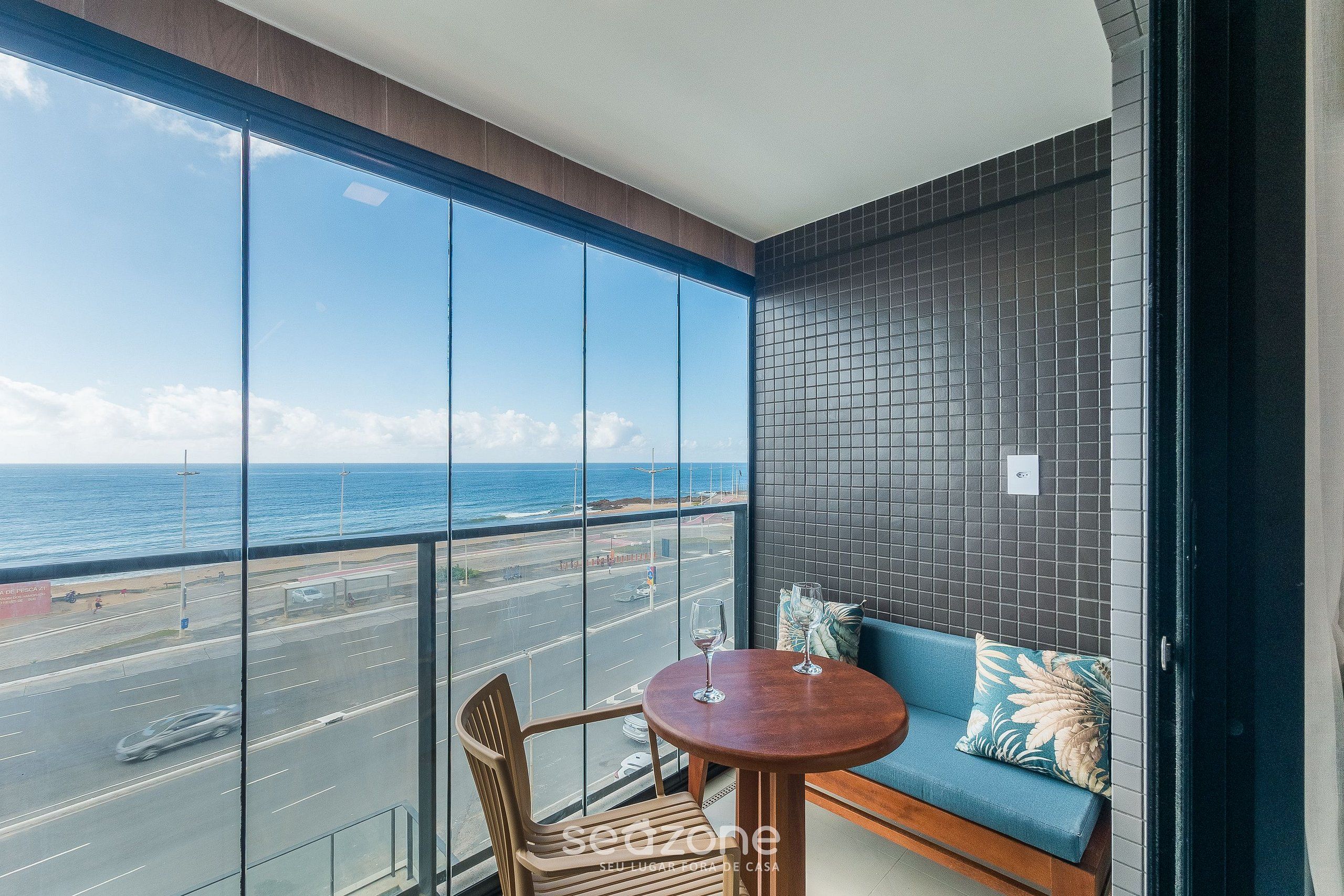 A balcony with a view of the ocean and a road, a round table with two chairs, and a blue sofa with a patterned pillow.