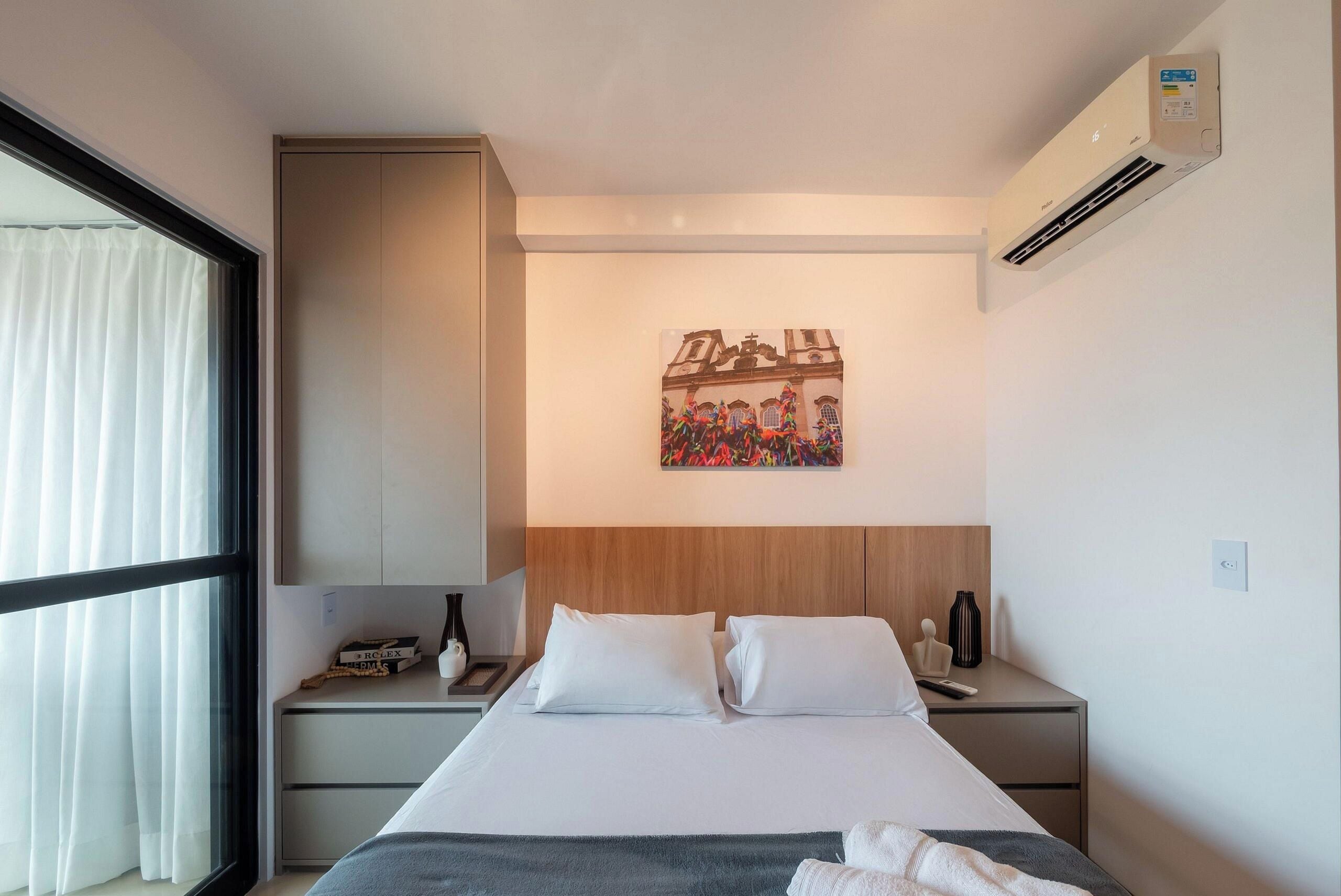 A hotel room with a bed, a nightstand, a window with curtains, a wall-mounted air conditioner, and a painting above the bed.