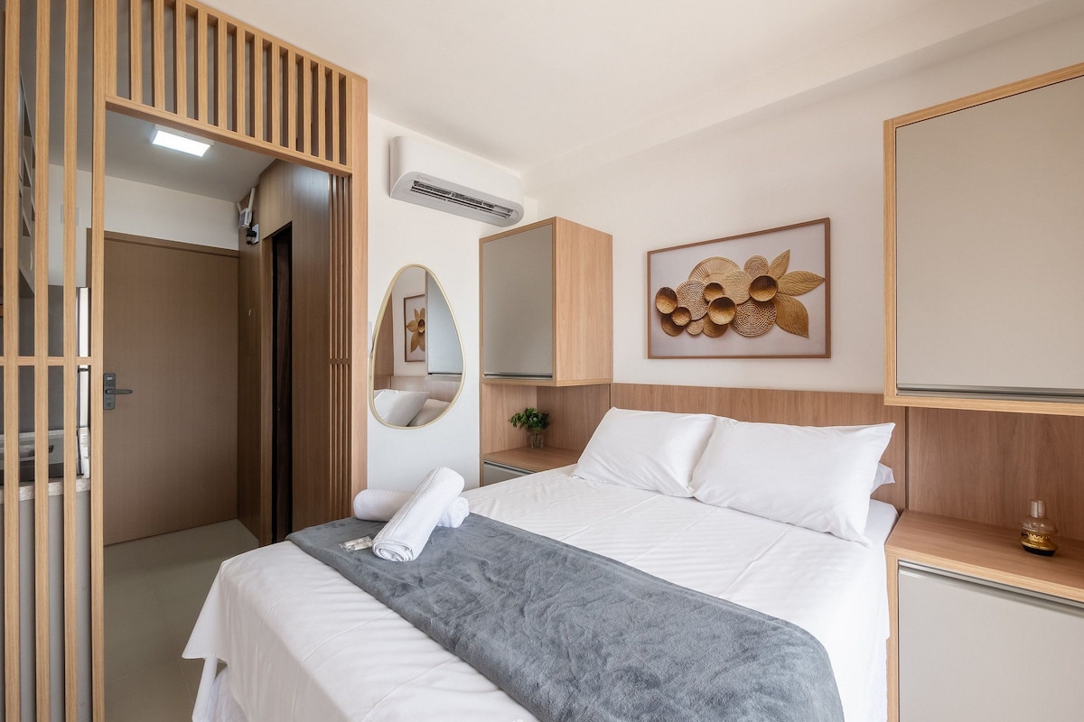A hotel room with a bed, a mirror, an air conditioner, a bedside table, and a wall-mounted artwork.