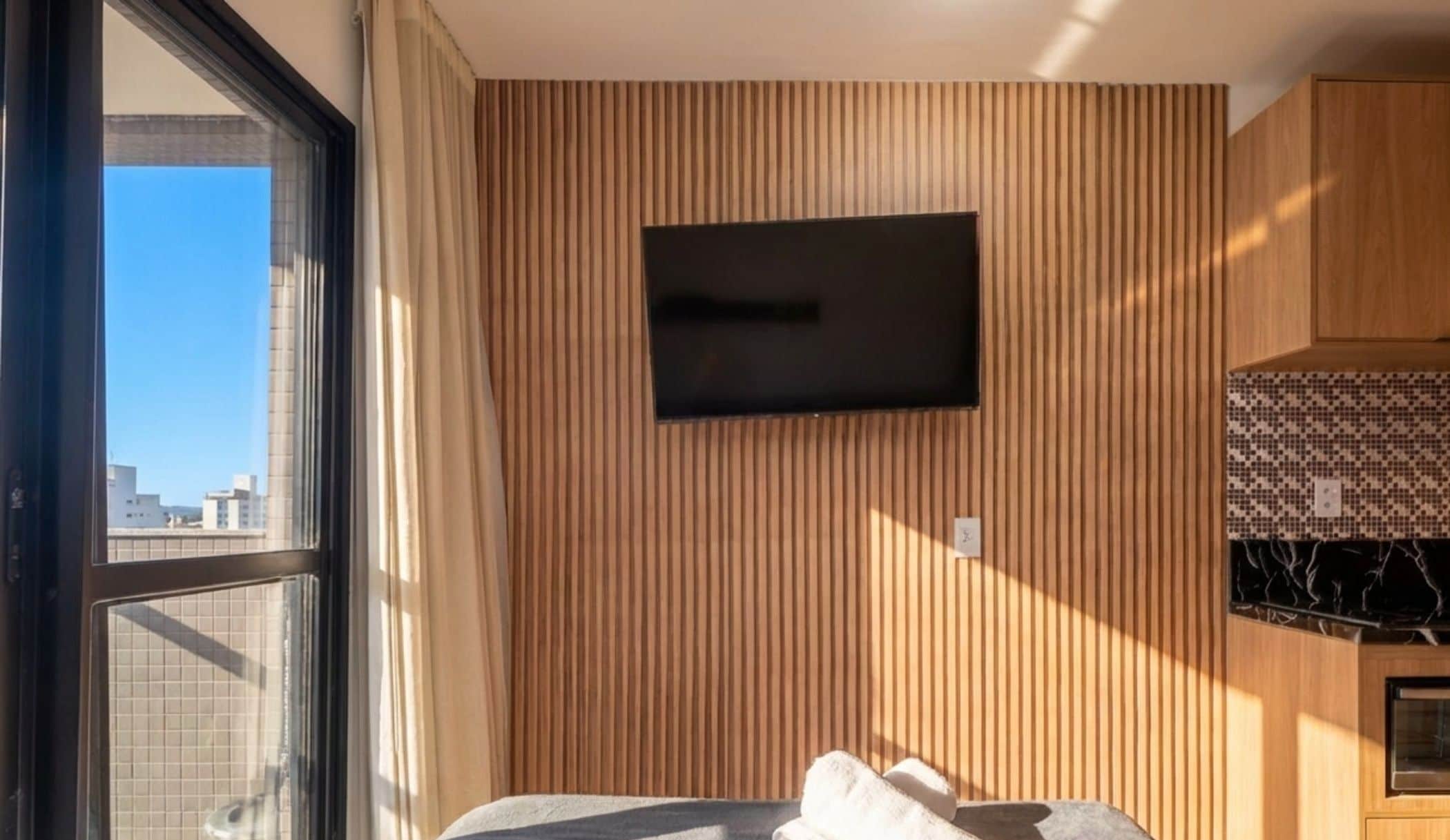 Una habitación con cama, televisión montada en la pared, ventana con cortinas y vistas a edificios.
