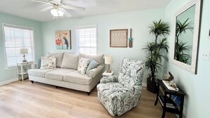 Ein Wohnzimmer mit Deckenventilator, einer Couch, einem gemusterten Sessel, einem kleinen Tisch mit Lampe und einer Topfpflanze.