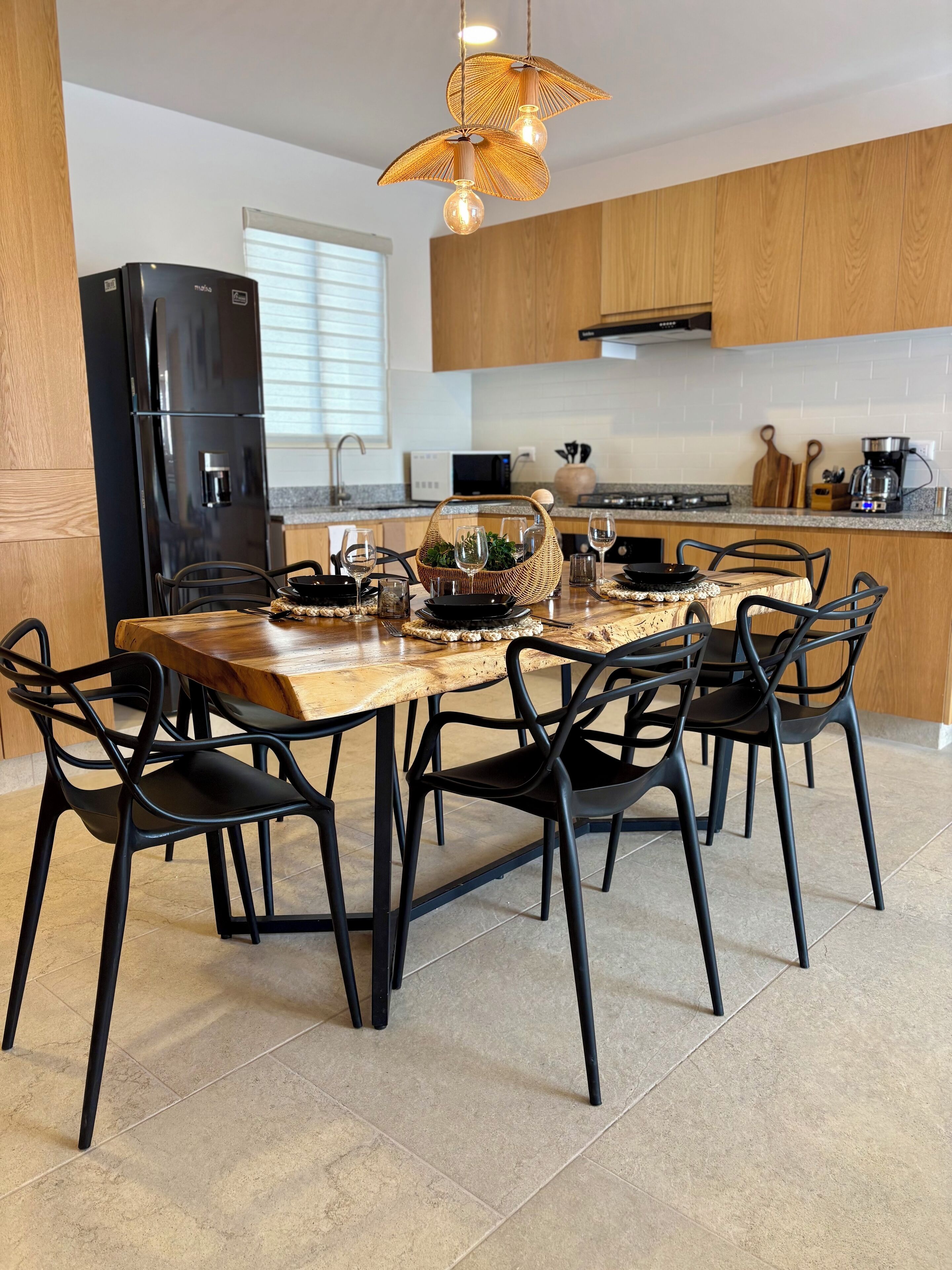 A modern kitchen with a wooden dining table, black chairs, a black refrigerator, and a hanging light fixture.
