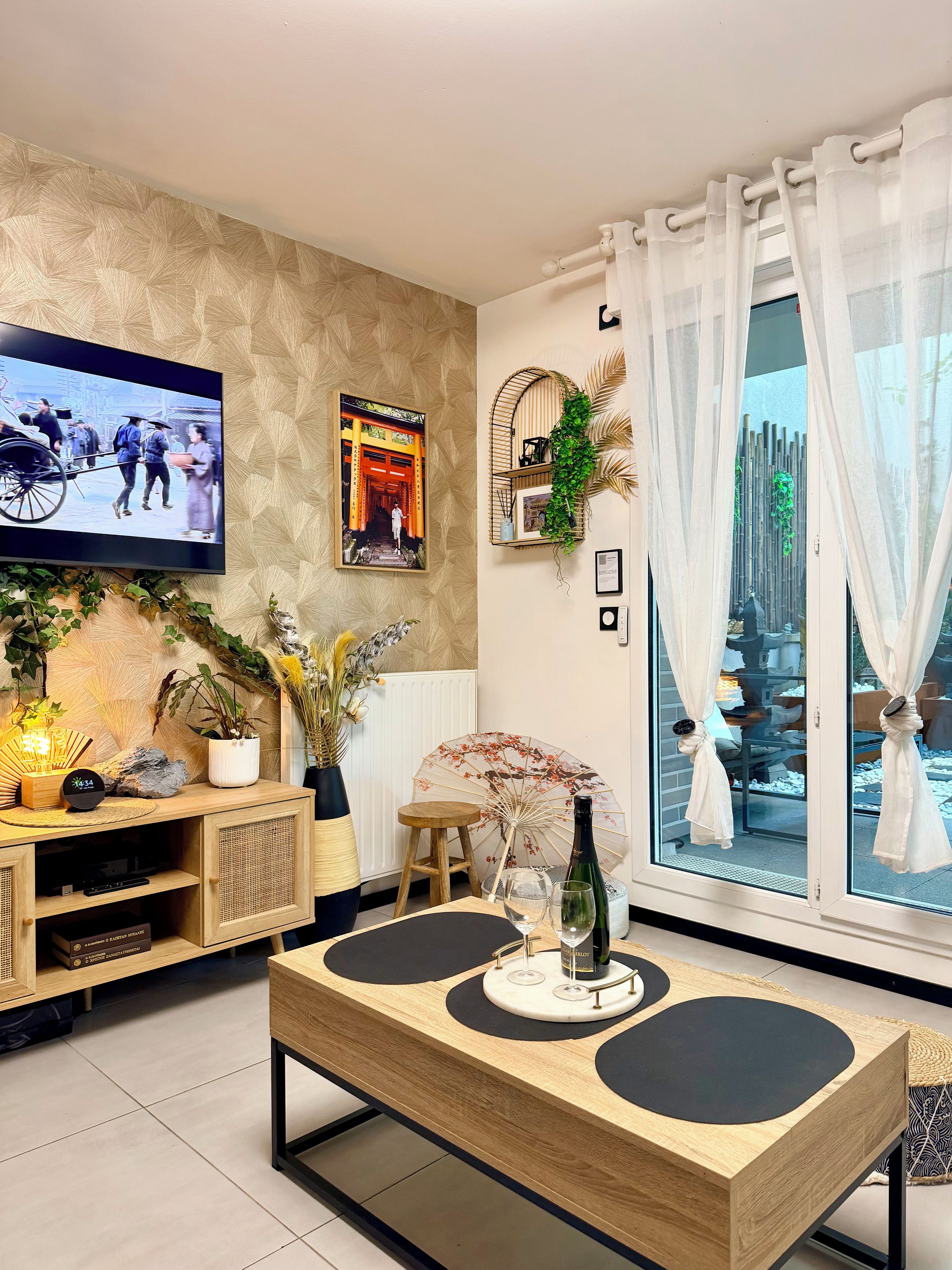 A modern living room with a TV, wooden furniture, a dining table with black mats, a potted plant, and a view of a bamboo fence through sheer curtains.