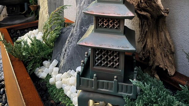 Une lanterne de style japonais traditionnel, en forme de pagode, se dresse dans un jardin aménagé, avec un grand rocher et une haie de bambous en arrière-plan.