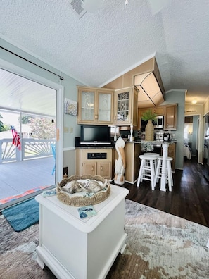 A spacious living area with a large window, a television, and a kitchen with wooden cabinets and a dining area. - 6010 Almost Heaven By The Sea (Myrtle Beach)