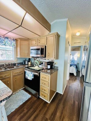 A kitchen with wooden cabinets, a stainless steel microwave, and a stove. There is a window with a curtain, and a rug on the floor. - 6010 Almost Heaven By The Sea (Myrtle Beach)