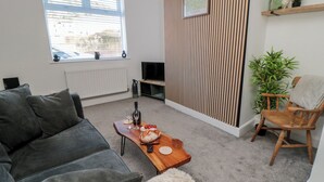 A living room with a grey sofa, a wooden coffee table with food and drink, a fluffy white rug, a wooden chair with a towel, a TV, and a radiator. - Islas Sea Forge (Saltburn-by-the-Sea)