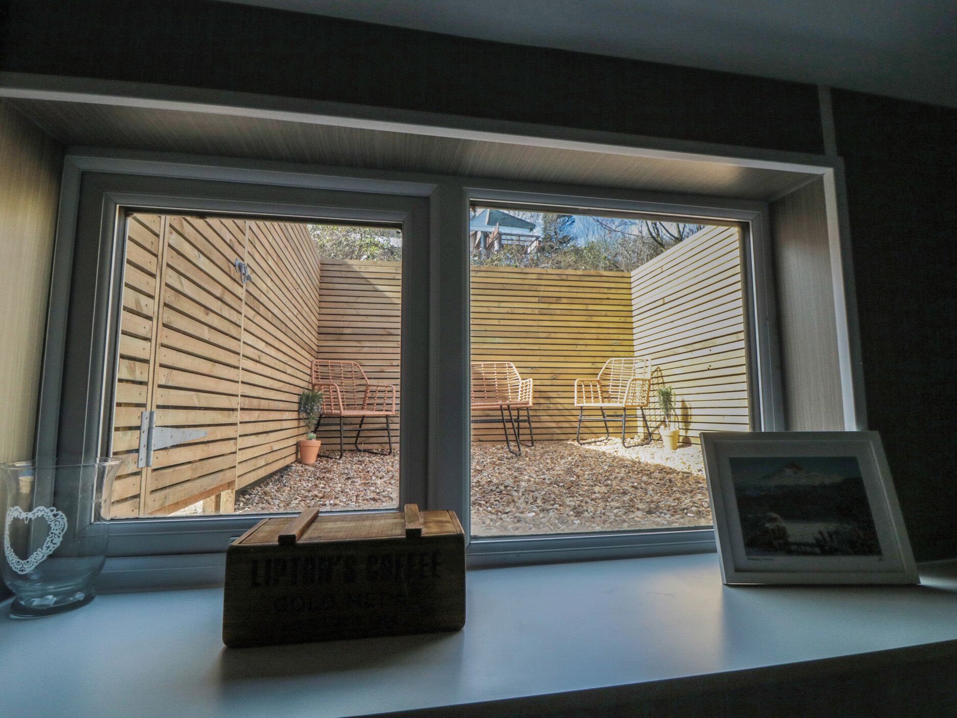 A window with a view of a wooden deck, two chairs, and a small table with a coffee box.