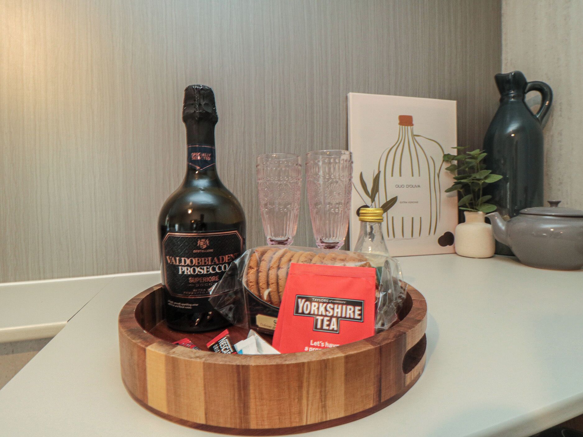 A wooden tray with a bottle of Valdobbiano Prosecco, two glasses, a bottle of Olio D'olio, a pack of Yorkshire Tea, and a small vase with a plant.