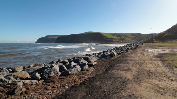 A coastal road with a rocky breakwater leading to the sea, a clear sky, and a hilly landscape in the background. - Islas Sea Forge (Saltburn-by-the-Sea)