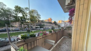 Un balcón con mesa y sillas, barandilla con plantas en macetas y vista a una calle con coches y edificios.