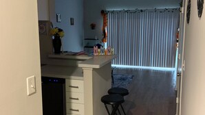 A kitchen with a central island, a dining area with a blue rug, and a living space with a curtain and a plant.