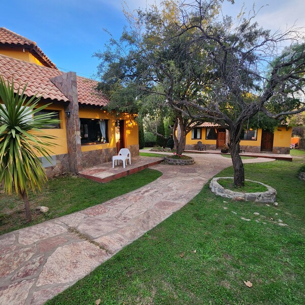 A courtyard with a pathway leading to a building with a tiled roof and stone walls. There are two white chairs on the patio, and a tree with yellow flowers in the center. - Merlo Trail Lodge (Merlo)