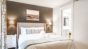 Een netjes opgemaakt bed met hoofdbord, twee nachtkastjes met lampen, een schilderij aan de muur, en uitzicht op een badkamer door een geopende deur.