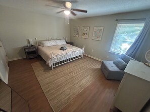 Une chambre à coucher comprenant un lit, un ventilateur de plafond, une table de chevet, une commode, une fenêtre avec des stores et des œuvres d’art murales.