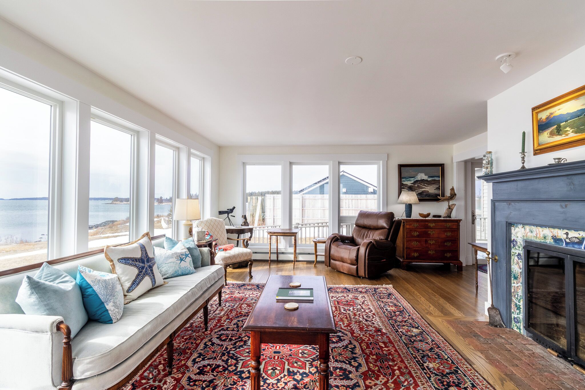 Un salon comprenant un canapé, un fauteuil et une cheminée, de grandes fenêtres offrant une vue sur la mer, et un tapis à motifs.