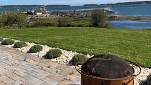 A rustic outdoor seating area with a metal pot stove, a paved walkway, and a view of a lake and pier. - OCEANFRONT New Custom Built 3Bdrm Handicap Accessible Home VIEWS & BEACH ACCESS (Jonesport)
