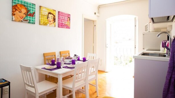 Ein Essbereich mit weißem Tisch und Stühlen, eine Küche mit Spüle und Schränken sowie ein helles Fenster.