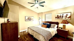 A bedroom with a bed, a nightstand, a desk, a ceiling fan, and wall art.