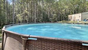 Una piscina con cerramiento de mimbre, rodeada de altos pinos y un edificio al fondo.