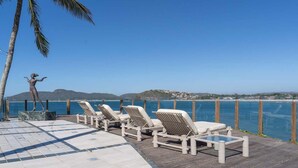 Eine Terrasse mit Liegestühlen mit Blick auf die Küste, eine Statue und einen klaren Ausblick auf das Meer und die fernen Hügel.