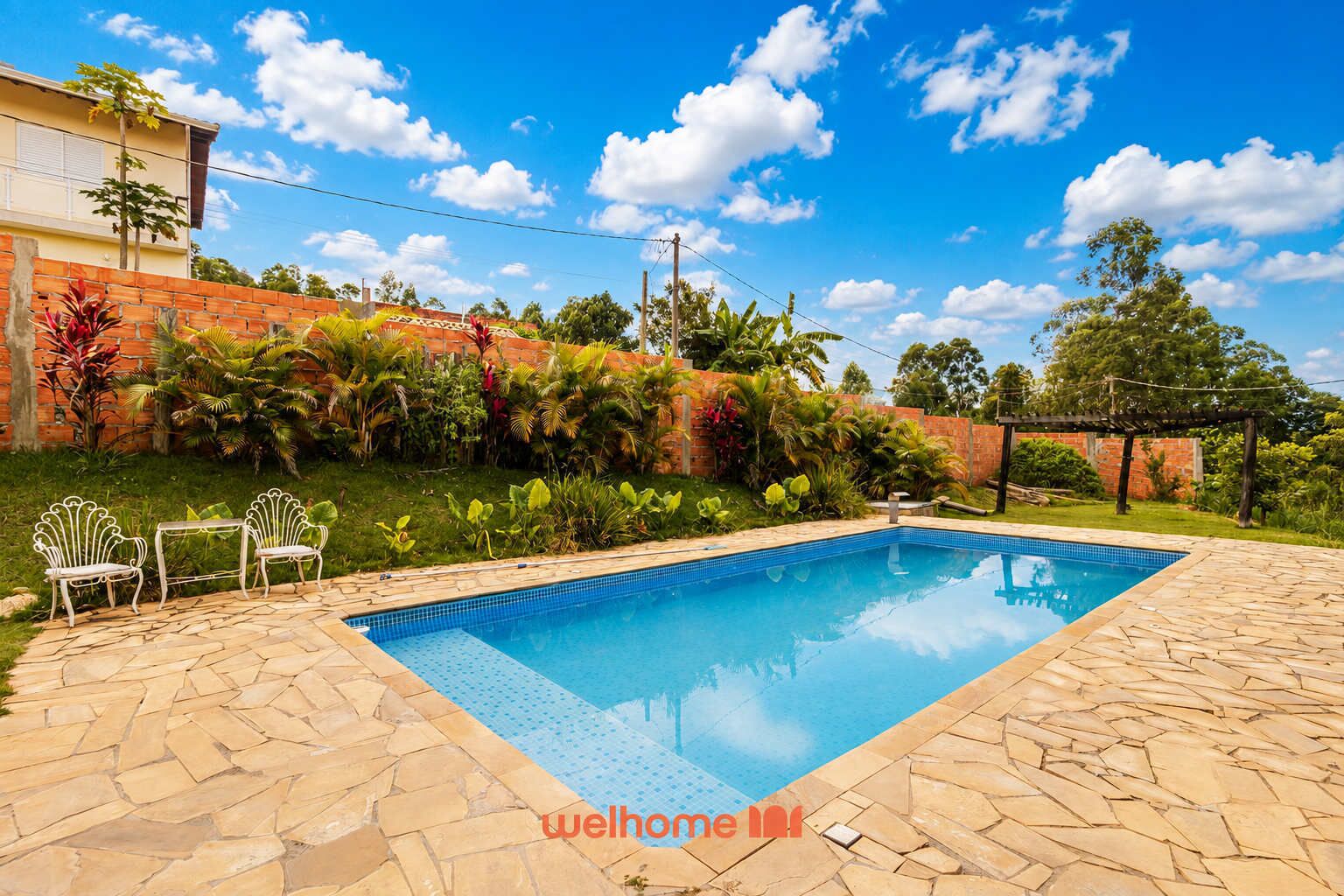 A rectangular swimming pool with a tiled edge, surrounded by a stone patio. There are two white metal chairs and a small table on the patio.